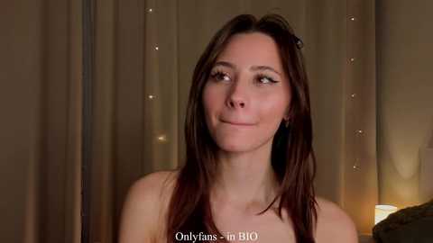 Media: Video of a fair-skinned woman with long brown hair, wearing minimal makeup, against a softly lit, beige curtain background.