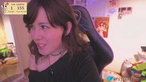 Video of a light-skinned woman with wavy brown hair, wearing a black top, sitting on a black gaming chair in a cluttered room. Background includes a TV, posters, and various toys.