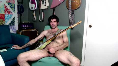 Video of a muscular, tattooed, naked man with short dark hair playing an upright bass in a messy room with guitars on the wall.