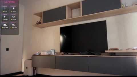 Video of a minimalist living room with a large black flat-screen TV mounted on a sleek, light wood TV stand. The background is a plain beige wall with a digital thermometer displaying indoor and outdoor temperatures.