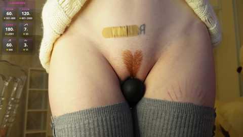 Video of a person with pale skin, wearing gray thigh-high socks, with a black anal plug and a beige bandage on their stomach, standing indoors.