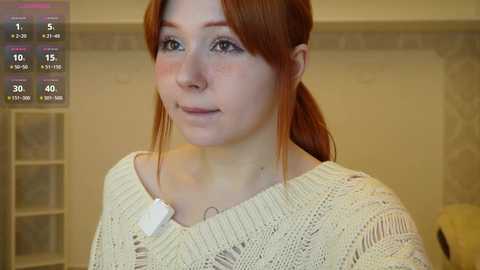 Video of a fair-skinned woman with freckles and red hair in a beige knitted sweater, set in a beige room.