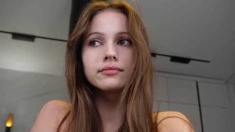 Video of a young, fair-skinned woman with long, wavy brown hair, wearing a light orange top, looking contemplative in a modern, minimalist room with white walls and a hanging light fixture.