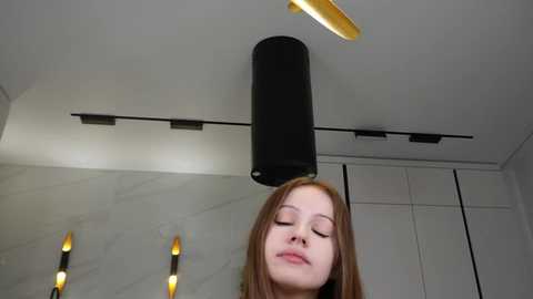 Video of a young woman with fair skin and long, straight red hair, eyes closed, standing in a modern, minimalist room with a white ceiling, black cylindrical light fixture, and sleek white cabinets.