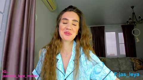 Video of a Caucasian woman with long brown hair, wearing a light blue robe, smiling with closed eyes, in a modern living room with beige walls, air conditioner, and windows.