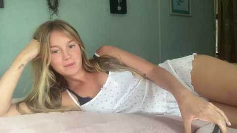 Video of a blonde, slender woman with fair skin, lying on a bed in white polka-dot pajamas, smiling, in a cozy, softly lit room.