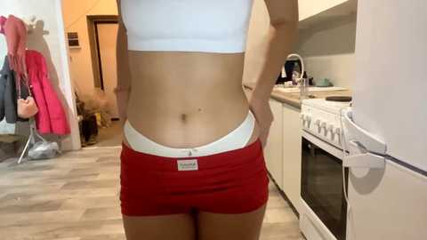 Video of a slender woman with light skin, wearing a white sports bra and red shorts, standing in a modern kitchen with white cabinets, wooden floor, and hanging coats.