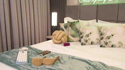 Video of a modern bedroom with a grey upholstered headboard, white sheets, and a turquoise blanket. Decor includes floral pillows, a round lamp, and a beige clutch.
