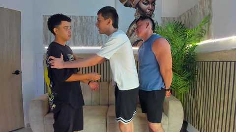 Video of three men in a living room: one restrained, another restraining him, and a third watching. They wear casual clothes. Background includes a couch, wall art, and plants.