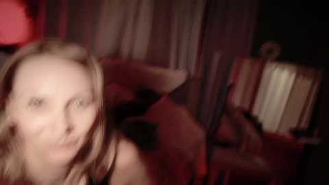 A blurred video shows a smiling woman with shoulder-length blonde hair, possibly in a dimly lit room with red lighting. In the background, indistinct figures are visible, suggesting a candid, possibly intimate setting.