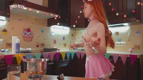 Video of a slender, fair-skinned woman with long red hair in a pink maid costume, mixing cake batter in a kitchen.
