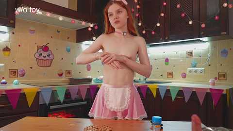 A video of a fair-skinned, young woman with red hair, wearing a pink maid outfit, covering her small breasts, standing in a colorful, cartoon-themed kitchen.