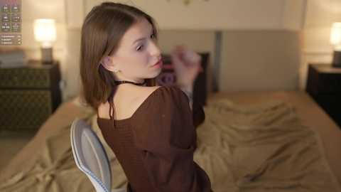 Video of a young woman with straight brown hair, wearing a brown off-shoulder top, sitting on a bed in a dimly lit bedroom with beige linens.