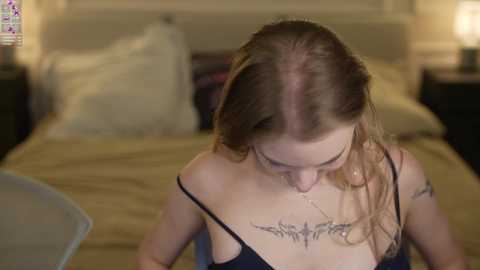 Video of a young woman with light skin, long brown hair, and a tribal tattoo on her chest, sitting on a bed in a dimly-lit bedroom.