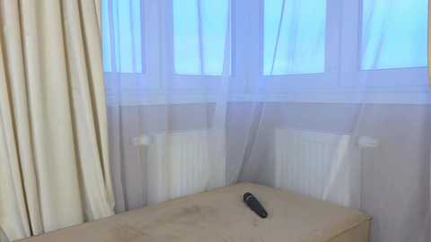 A video of a minimalist bedroom with a beige bedspread, sheer white curtains, and a black remote control on the bed. The room has soft, diffused natural light filtering through the windows.
