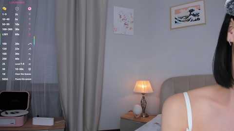 Video of a woman in a white camisole, facing away, in a minimalist bedroom with beige walls, a bed, a lamp, and a window with sheer curtains.