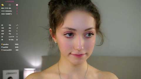 Video of a young woman with fair skin, brown hair pulled back, wearing a necklace. Background shows a calendar, and blurred furniture.