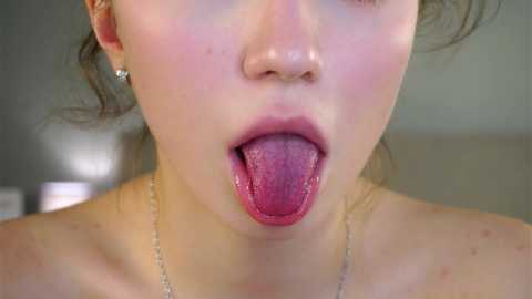 Close-up video of a fair-skinned woman with light brown hair, wearing a gold necklace, tongue sticking out, and light makeup. The background is blurred, featuring a neutral-toned indoor setting.