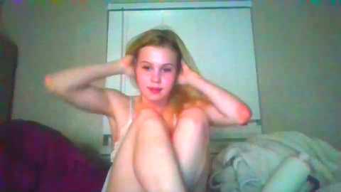 Video of a fair-skinned, blonde woman in a white bra, sitting on a bed with rumpled sheets, adjusting her hair.