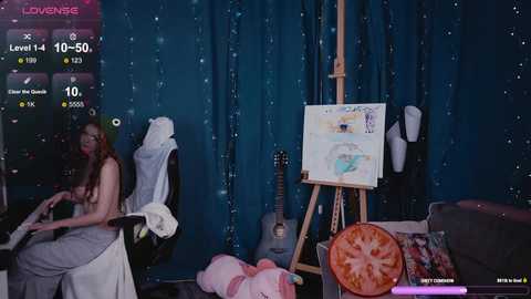 A video of a young woman with long hair painting in a dimly lit room with a blue backdrop, surrounded by various art supplies, a guitar, and a TV screen displaying a \"Love Live!\" game.