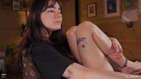 A video of a young woman with dark hair and a tattoo on her thigh, sitting on a couch in a cozy, wooden-paneled room with art on the walls.