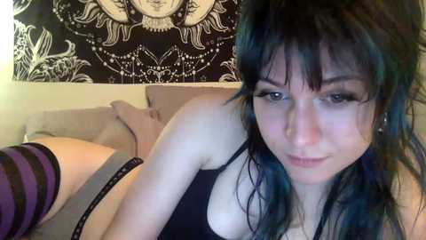 A video of a young woman with pale skin, blue hair, and dark makeup, lying on a bed, wearing a black tank top and striped thigh-high socks, in a dimly lit room with a black tapestry on the wall.