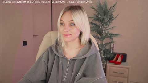 Video of a young, fair-skinned woman with platinum blonde hair, wearing a gray hoodie, sitting in a beige chair in a minimalist room with a potted plant and a pair of red high heels on a side table.