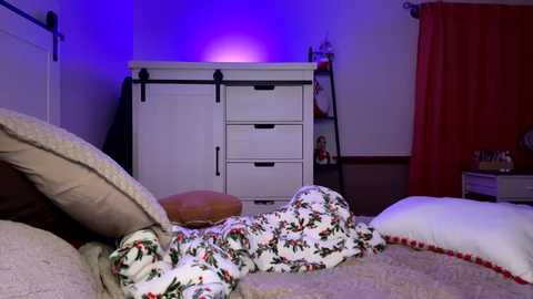 Video of a cozy bedroom with a white dresser, floral blanket, beige pillows, red curtains, and purple accent lighting.
