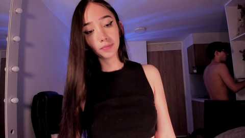Video of a young Asian woman with long black hair, wearing a sleeveless black top, standing in a dimly lit bedroom with a mirror and a man in the background.
