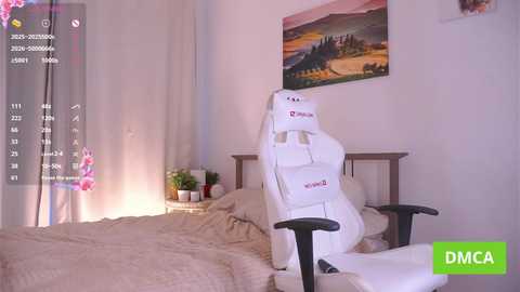 Video of a minimalist bedroom with a white gaming chair, beige bedding, a potted plant, and a sunset landscape painting on the wall.