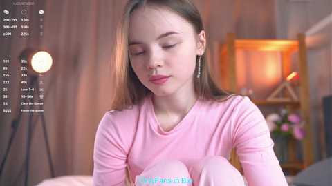 Video of a young, fair-skinned girl with straight brown hair, wearing a pink long-sleeve shirt, holding a pink plush toy, in a cozy, warmly lit room with a bookshelf in the background.