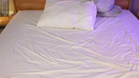 Video of a neatly made bed with white sheets, pillows, and a wooden headboard, bathed in soft purple light.