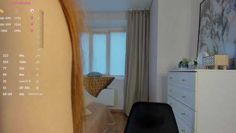 Media: Video of a modern, minimalist bedroom with beige walls, a white dresser, and a black chair; a bed with a woven blanket, beige curtains, and a floral painting on the wall.