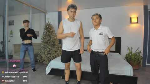 Video of three young men: one tall, muscular, wearing black shorts and a white tank top; another shorter, wearing a white polo and black pants; a third, standing by a bed with a Christmas tree in a modern room.