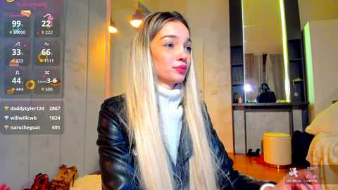 Video of a young woman with long, straight blonde hair and fair skin, wearing a black leather jacket and white turtleneck sweater, seated in a modern living room with a digital screen displaying weather information and social media notifications.