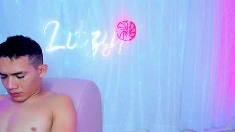 Video of a shirtless, Asian man with short hair, wearing a gold chain, sitting on a white chair. Neon \"Luvly\" sign and pink swirl on a light blue, textured background.