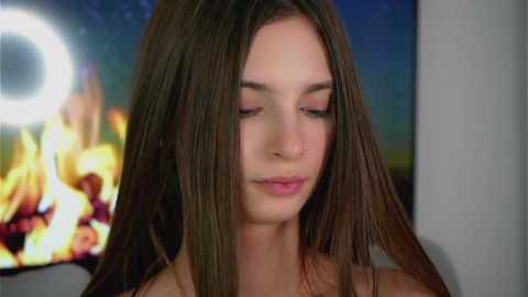Video of a young woman with long, straight brown hair, fair skin, and closed eyes, standing in front of a burning fireplace in a cozy room.