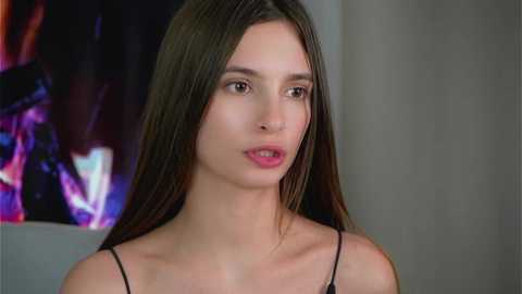 Video of a young woman with long brown hair, fair skin, and a slim physique, wearing a black spaghetti strap top, standing against a blurred, colorful abstract background.