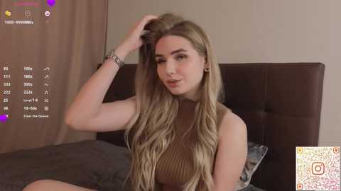Video of a young, light-skinned woman with long, wavy blonde hair, wearing a sleeveless brown top, sitting on a bed. The room has beige walls and a dark headboard.