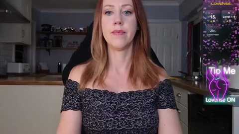 Video of a fair-skinned, red-haired woman in a black lace dress, sitting in a modern kitchen with a digital screen displaying \"Tip Me\" and \"Lovense ON.\