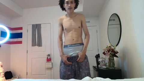 Video of a young, shirtless, skinny man with curly hair in plaid pajama pants standing in a dimly lit, cluttered bedroom.