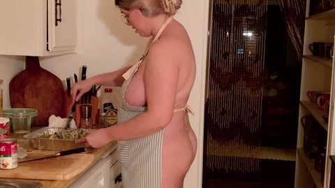 Video of a middle-aged woman with a fair complexion, short blonde hair, and average build, wearing a striped apron, preparing food in a kitchen. The background features a wooden chopping board, utensils, and various cooking items.