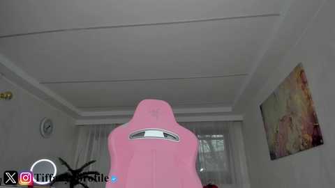A video of a pink gaming chair in a well-lit room with a white ceiling and a large window adorned with sheer white curtains. The room features a colorful abstract painting on the right wall.