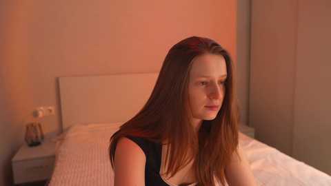 Video of a young woman with long, straight brown hair, wearing a black tank top, sitting on a bed with white linens in a softly lit, minimalist bedroom.