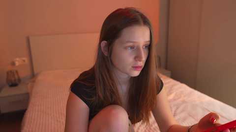 A video of a young, fair-skinned woman with long brown hair, wearing a black top, sitting on a bed in a dimly lit, modern bedroom.