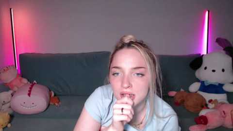 A video shows a young woman with fair skin and blonde hair, wearing a blue t-shirt, licking a lollipop in a room with a grey couch, stuffed animals, and pink LED lights.