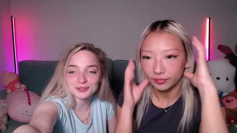Video of two young women, one blonde and one with long, straight hair, posing with hands in front of a gray couch, neon lights in background.