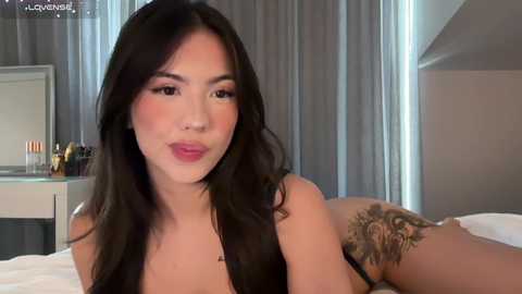 A video of an East Asian woman with long, straight black hair and light skin, wearing black lingerie, lying on a bed in a modern bedroom with gray curtains. She has a tattoo on her thigh.