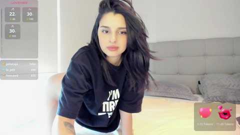 A video of a young woman with long black hair and fair skin, wearing a black t-shirt, sitting on a bed in a modern, minimalistic bedroom.