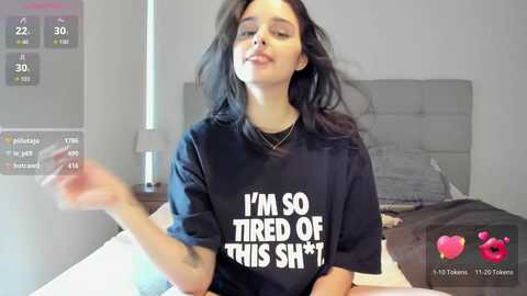 Video of a woman with long, dark hair, wearing a black t-shirt with \"I'M SO TIRED OF THIS SHIT\" in white letters. She is in a modern bedroom, lying on a bed with a gray headboard.